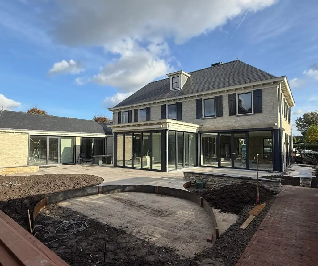 Two-story brick house with large glass doors and black shutters under construction; foreground shows dug soil and curved brick edging for landscaping.